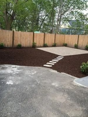Backyard patio with stone path, mulch beds, wooden fence, and trees behind concrete driveway
