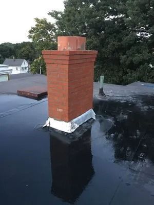 Brick chimney on a flat black roof with trees and houses in the background