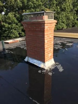 Brick chimney on a flat roof with metal cap and dark tar flashing, surrounded by trees