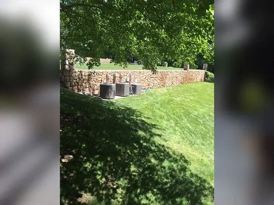 Grassy hillside with trees, a stone wall, and two trash bins in a sunny park area