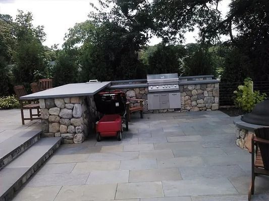 Stone outdoor patio with steps, built-in grill area, and red lawn equipment under a small roof