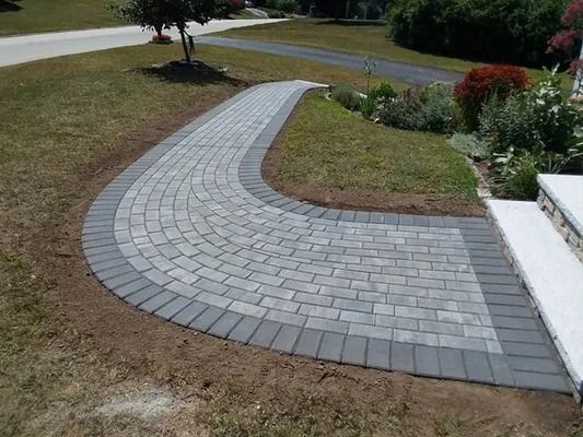 Curved gray paver walkway through a grassy yard beside a white house and garden beds