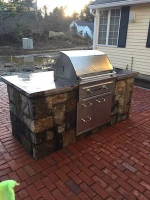 Outdoor built-in stainless steel grill with stone base on a brick patio beside a house.