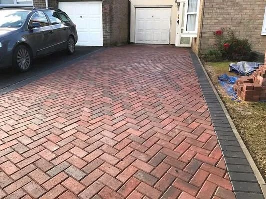 Brick driveway between a parked car and garage doors, bordered by a curb and grass with debris on the right.