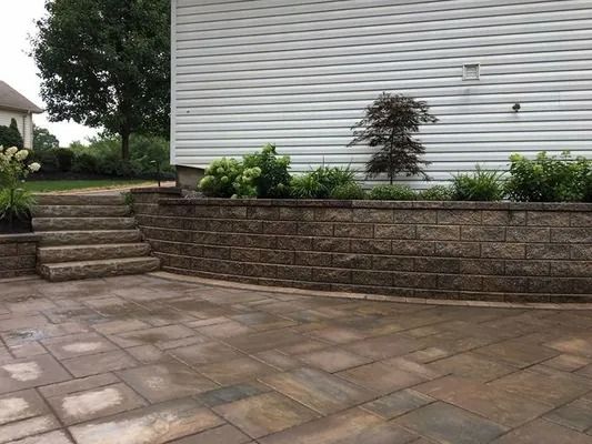 Stone patio with steps, retaining wall, and white house siding with shrubs along the base.