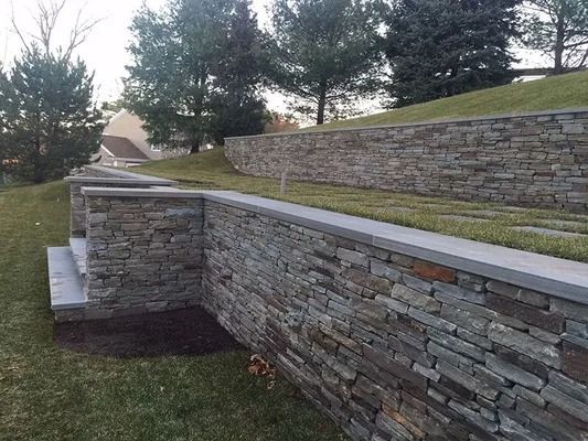 Stone retaining wall along a landscaped hillside with grass and trees under an overcast sky