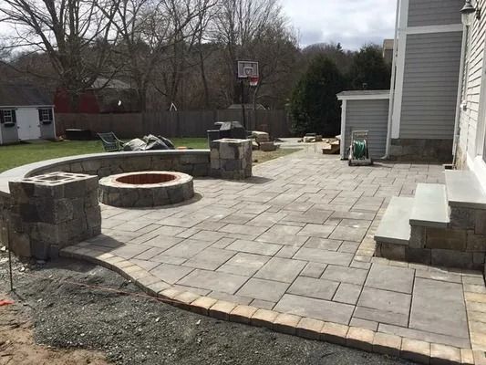 Stone patio with a circular fire pit and steps beside a gray house in a backyard