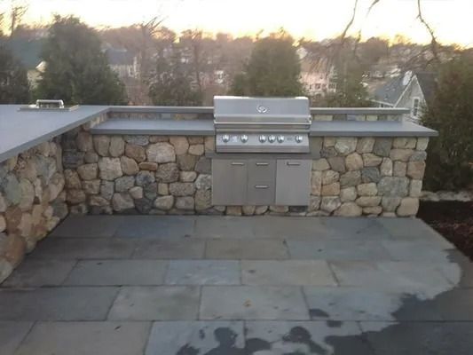 Outdoor stone barbecue grill on a patio at dusk, with a stone wall and houses in the background