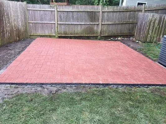 Red paved patio area in a backyard with a wooden fence and grass border