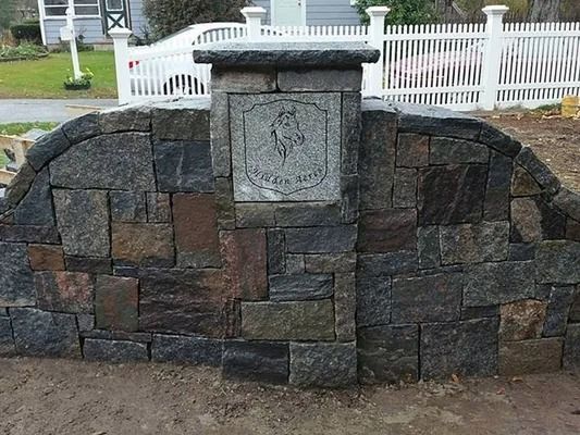 Stone memorial wall with a carved plaque in front of a white fence and sidewalk