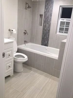 Modern bathroom with a white tub, toilet, vanity, gray tile, and window.