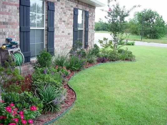 House front with flower bed, shrubs, and green lawn beside brick siding and windows