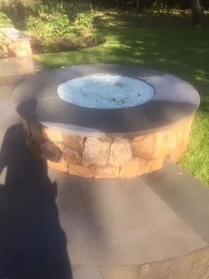 Outdoor stone fire pit with a circular metal rim on a concrete patio, surrounded by grass and sunlight