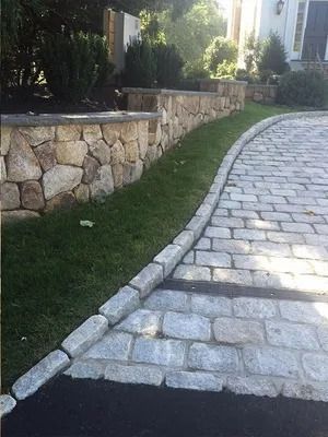 Curved stone-paved walkway beside a grassy strip and stone retaining wall in a landscaped area