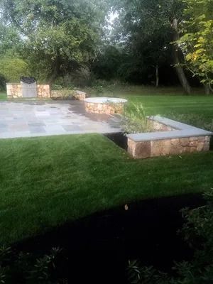 Stone patio and low retaining walls beside a green lawn and wooded garden area.