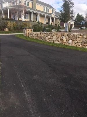 Residential street with black pavement, stone retaining wall, and large house with porch and driveway
