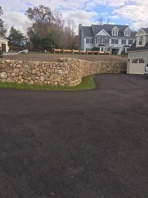 Stone retaining wall in front of a large gray house with a dark driveway and overcast sky.