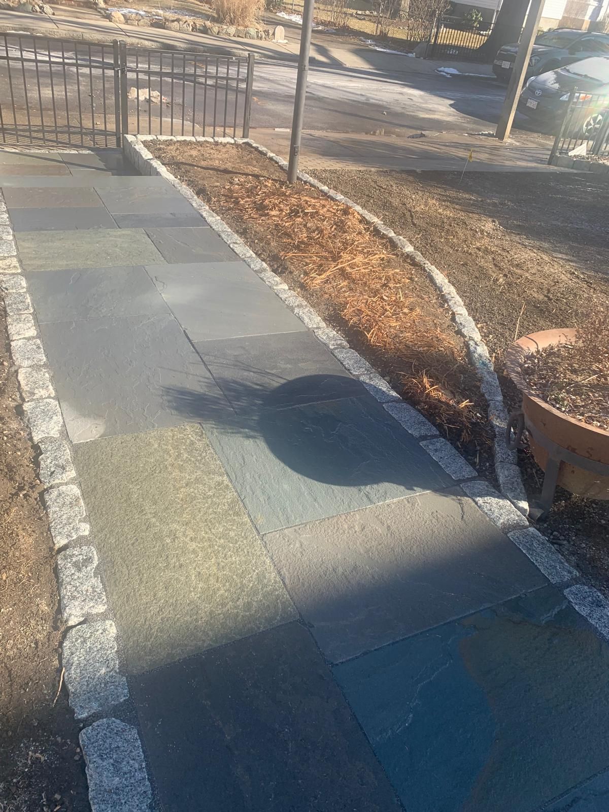 Sunlit sidewalk with patchwork pavement, curb, leaf-lined edge, and a fence casting shadows.