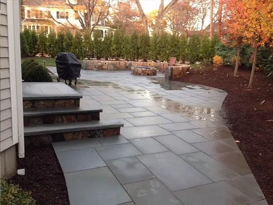 Backyard patio with wet stone pavers, steps, and a covered grill beside landscaping under warm light