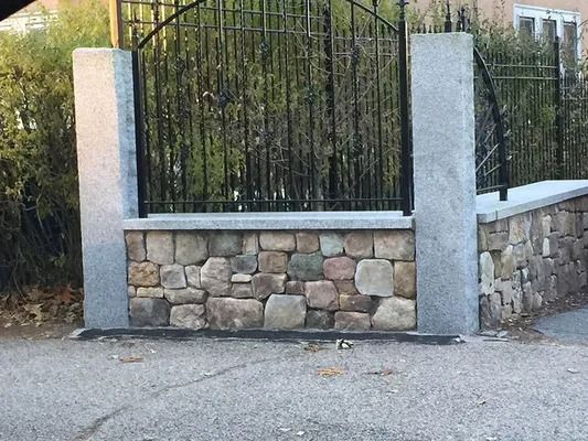 Stone-and-concrete fence with black metal gate along a roadside.