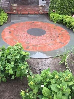 Red brick circular patio with a dark center, bordered by gray stone and green garden plants.