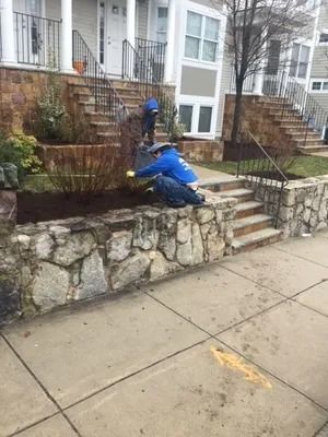 Worker in blue jacket trimming shrubs along a stone wall beside apartment steps and sidewalk.