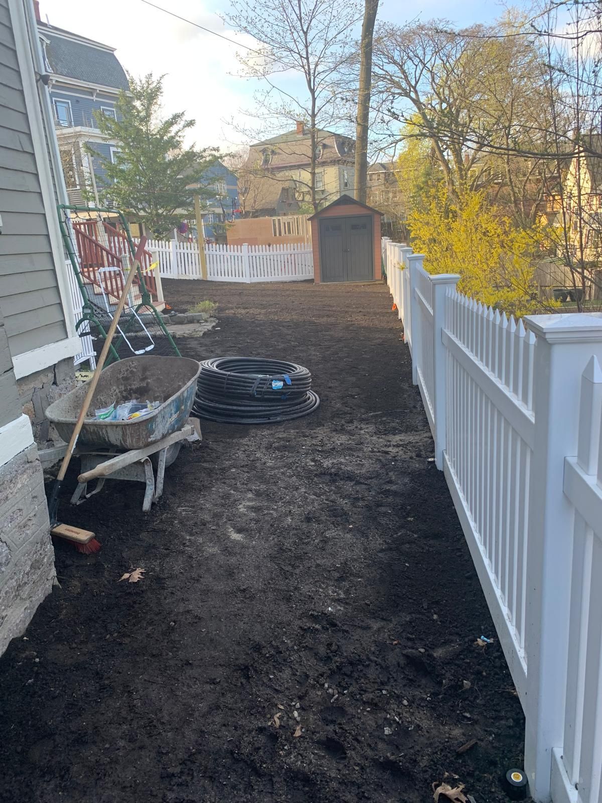 Backyard with fresh dirt, a white fence, and garden tools beside a house