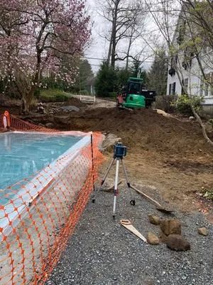 Construction site beside a swimming pool, with a green excavator on a dirt slope and survey tripod in foreground