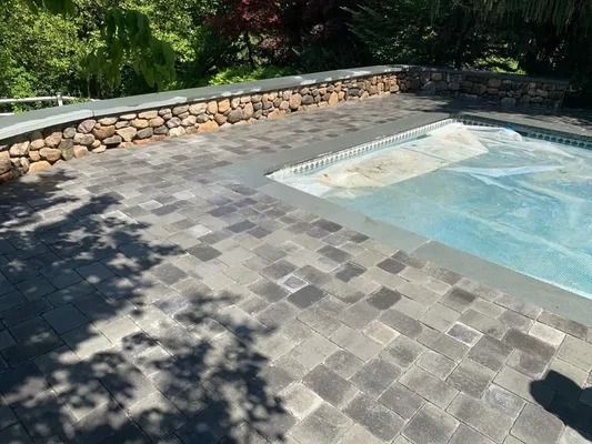 Stone pool deck beside a blue swimming pool, with a low rock wall and tree shadows.