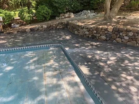 Stone-edged swimming pool beside a rocky wall and shaded trees in a sunny yard