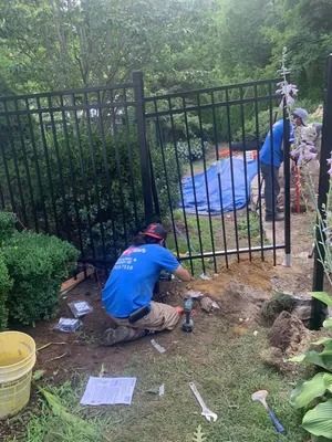 Worker kneeling by a black metal gate, using a tool near the ground in a muddy garden area