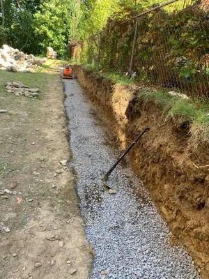 Gravel-filled trench beside a dirt path, with a fence and an orange wheelbarrow in the background