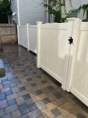 White vinyl fence along a wet brick-paved side yard beside a gray house
