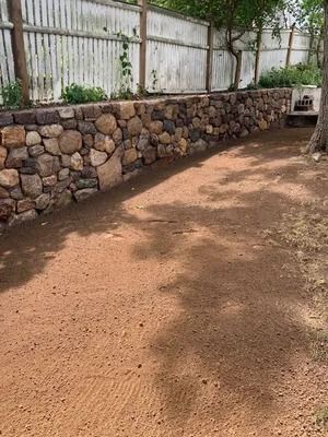 Dirt courtyard beside a stone retaining wall and white fence, with a tree casting shade.