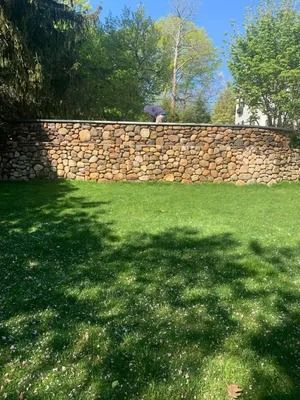 Stone retaining wall behind a sunlit grassy lawn with trees and blue sky overhead