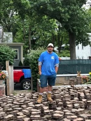 Worker standing on stacked pavers in a yard, wearing a blue shirt and cap, with a red pickup in the background