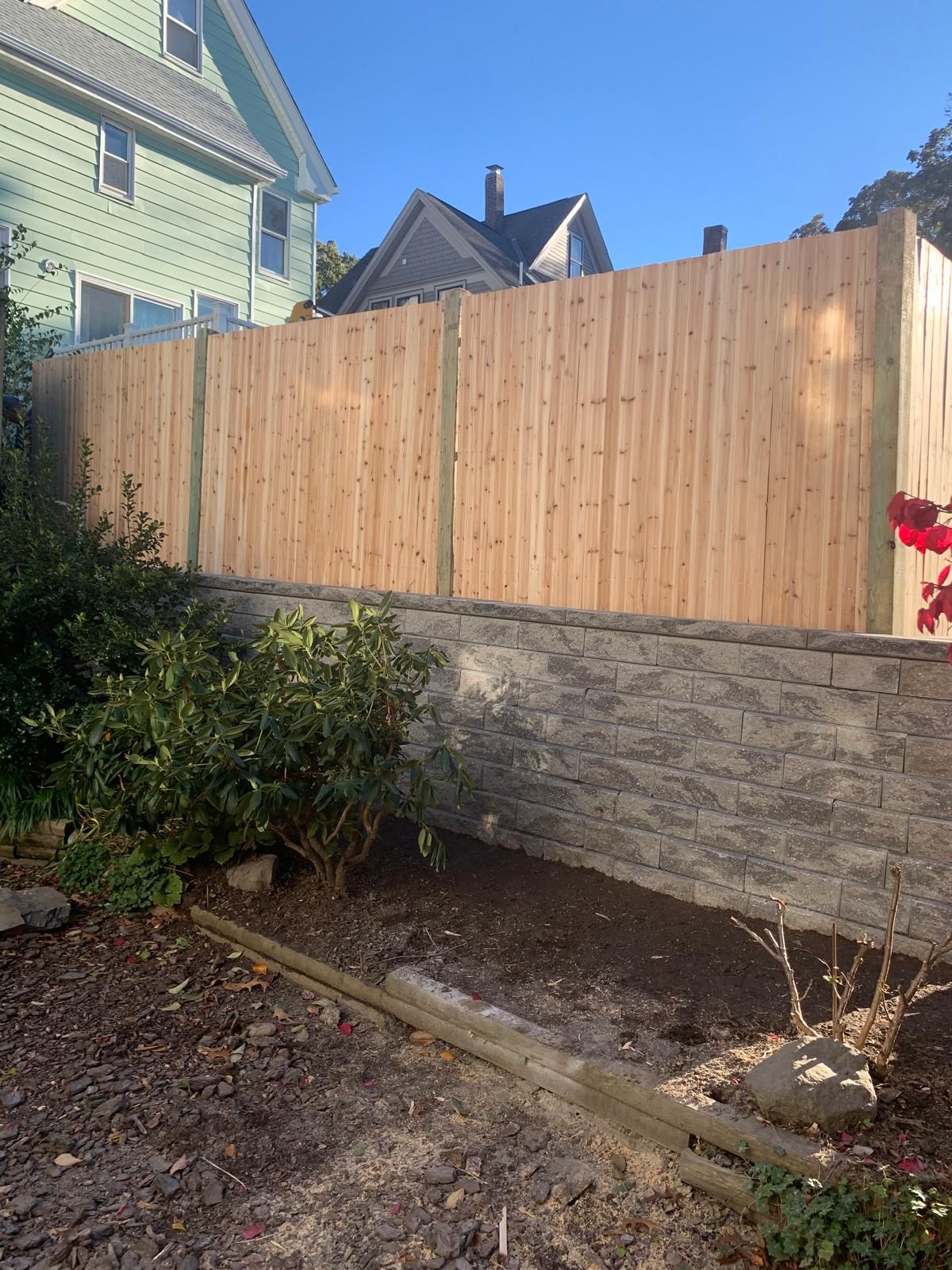Wooden fence above a stone retaining wall in a backyard garden, with a house and shrubs beside it.