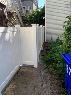 Narrow muddy side yard between white fence and house, with blue recycling bin on the right.