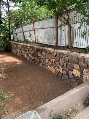 Stone retaining wall beside a dirt path, with a white fence and green vines along a sloped roadside.
