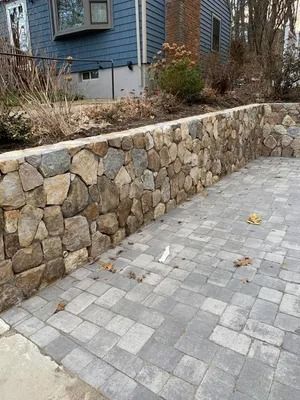 Stone retaining wall beside a gray paver driveway next to a blue house