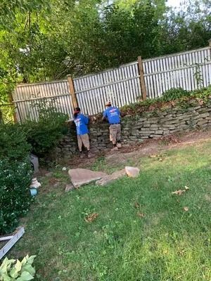 Two people in blue shirts work beside a stone wall and white fence in a grassy backyard.