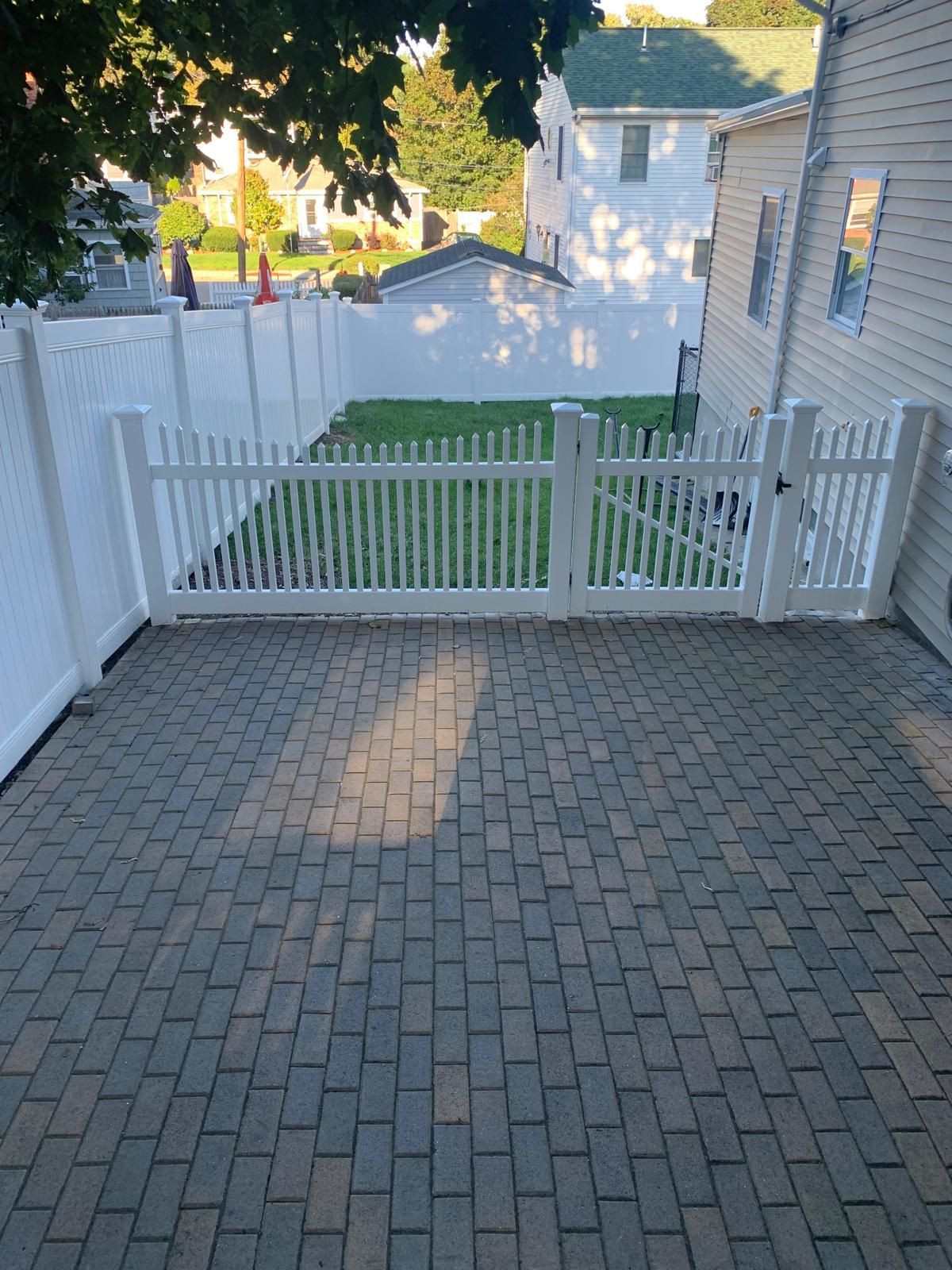 Brick patio facing a white picket gate and fence, with a grassy yard and houses beyond.