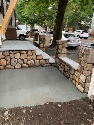Stone ramp and steps leading to a porch beside a tree-lined residential sidewalk with parked cars
