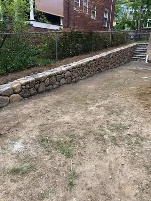 Stone retaining wall beside a dirt courtyard, with brick building and steps in the background.