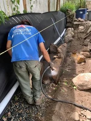Worker installing black pipe beside a stone trench, using a hose near a building wall.