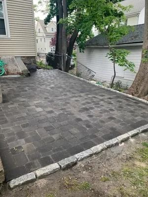 Freshly paved dark gray brick patio beside a house and garage, bordered by a tree and concrete edges