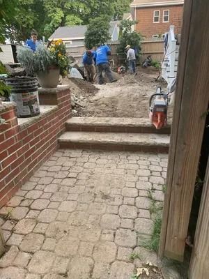 Workers digging up a brick patio and steps in a backyard, with equipment and dirt piled near the house.
