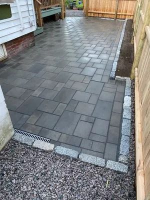 Freshly paved gray patio walkway beside a house and wooden fence, with gravel border and drain grate.