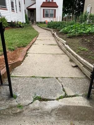 Cracked concrete walkway leading to a house entrance with a red awning and garden beds.