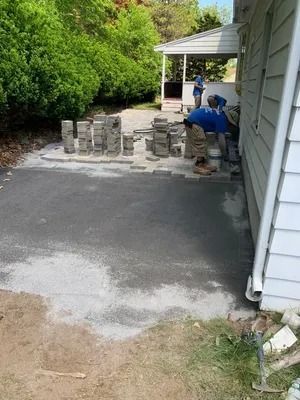 Worker laying stone pavers beside a house driveway with stacked materials and a porch in the background